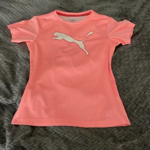 Rosa sportig t-shirt från Puma - Snygg rosa t-shirt från Puma med klassisk logga framtill. T-shirten är gjord i ett lätt och ventilerande syntetmaterial som håller dig torr. Perfekt för träning eller sportiga dagar. Kortärmad och figurnära passform för en aktiv look.