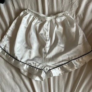Vita satinskjorts med svart kant - Snygga vita shorts i glansigt satinmaterial med elastisk midja och svart kant längs bensluten. Perfekta för en chill vibe eller som pyjamasshorts. Lätta och luftiga med en loose passform.