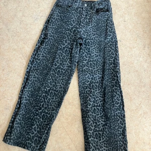 Leopardmönstrade wide jeans - Säljer ett par gråa jeans med svart leopardmönster. Byxorna har en rak och lite vidare passform, klassisk femficksmodell och hällor för bälte. Materialet är jeans och de har knapp och dragkedja framtill. Perfekta för dig som vill sticka ut med ett djurmönster.