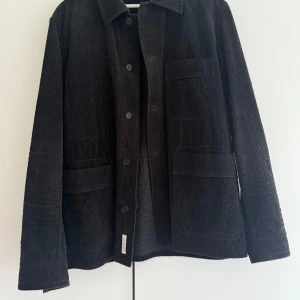 Svart manchester overshirt med fickor - Säljer en svart overshirt i manchester med klassisk krage och stora fickor framtill. Jackan har knäppning med knappar och raka ärmar. Köparen står för frakten. 