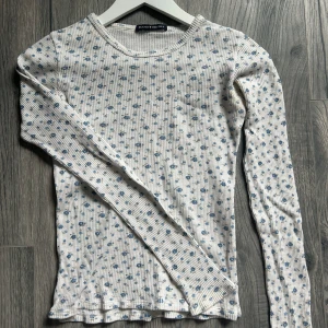 Vit blommig långärmad tröja Brandy Melville - Supersöt långärmad tröja från Brandy Melville i vit med små blå blommor över hela. Tröjan är ribbad och har en tight passform med rund halsringning. Perfekt att styla med jeans eller kjol för en chill look.