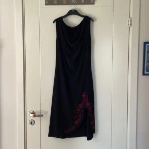 Svart klänning med röd brodyr Ette Lou - Svart ärmlös långklänning från Ette Lou med snygg draperad halsringning och röd broderad blomdetalj vid slitsen. Klänningen har en elegant och flowy passform som ger en cool och stilren look.