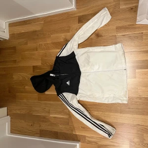 Adidas svartvit vindjacka med huva - Svartvit vindjacka från Adidas med klassiska tre ränder längs ärmarna och logga på bröstet. Jackan har huva, hel dragkedja och är tillverkad i lätt polyester. Perfekt för dig som gillar sportig stil och vill ha något som sticker ut.