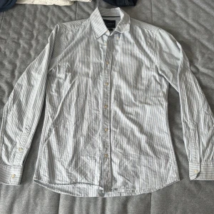 Randig skjorta från Bondelid, S - Snygg skjorta från Bondelid i vitt och ljusblått med smala ränder. Klassisk krage, långa ärmar och knappar framtill. Skräddarsydd passform (tailored fit) som sitter riktigt bra. Perfekt för dig som gillar stilrena och fräscha plagg.