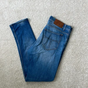Blå jeans från Tiger of Sweden - Snygga blå jeans från Tiger of Sweden med klassisk femficksdesign och ljus tvätt. Jeansen har normal passform, raka ben och detaljer i brunt läder vid linningen. Perfekta för dig som gillar stilrena och tidlösa jeans.