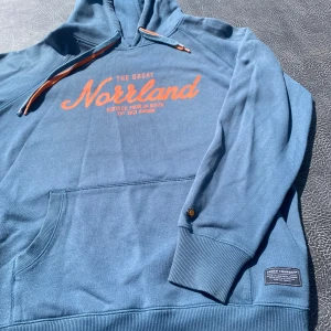 Mörk blå/grön hoodie från North Company M - Säljer en Mörk blå/grön hoodie från North Company med orange tryck 'The Great Norrland' på bröstet. Tröjan har huva med dragsko i blått och orange, känguruficka och ribbade muddar. Perfekt för dig som gillar snygga och bekväma plagg med streetkänsla. ( bilden på hoodien är inge bra för färgen är mycket mer mörk blå/grön i verkligheten)