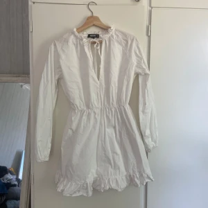 Vit kort klänning med volang - Supersöt vit kort klänning från Missguided med långa ärmar, volangkant nertill och knytning vid halsen. Resår i midjan ger en snygg passform och den har en lätt rynkad look. Perfekt för sommarens alla tillfällen!