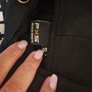 Svart hoodie från PXS Active Sports - Svart hoodie från PXS Active Sports med vit text på bröstet: 'Performance Athletic Team Champions Springfield'. Tröjan har huva med snörning och en stor magficka. Perfekt för dig som gillar sportig och avslappnad stil.