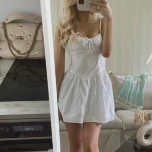 Vit kort sommarklänning med broderi - Supersöt vit kort klänning med tunna axelband och broderade blommor. Klänningen har en feminin tight passform med markerad midja, lätt  knytning framtill. Romantisk.