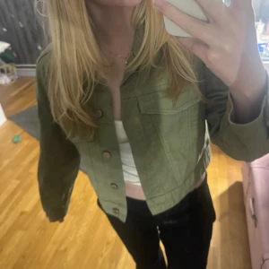 Olivgrön croppad jeansjacka Gina Tricot - Snygg croppad jeansjacka från Gina Tricot i olivgrönt. Klassisk krage, bröstficka med lock och knappar framtill. Perfekt till lager-på-lager och funkar till allt. Jackan har långa ärmar och är i mjuk bomull.
