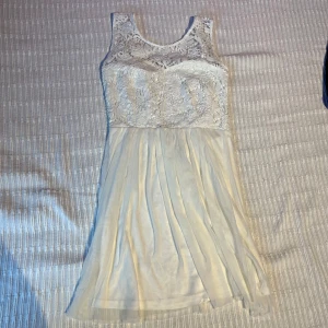 Vit spetsklänning med dragkedja - Superfin vit kortklänning med spets upptill och flowy kjol i tunt tyg. Klänningen är ärmlös och har en snygg dragkedja i ryggen. Perfekt för student eller sommarens alla tillfällen. Söt och romantisk look med blommig spetsdetalj.