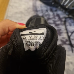 Svarta Nike Air sneakers med röda detaljer - Snygga svarta Nike Air sneakers med röda Swoosh-loggor och detaljer. Skorna har högt skaft, snörning och en mix av mesh och skinn för både stil och komfort. Sulan är svart med röda partier undertill och tydlig Nike Air-märkning på hälen.