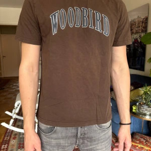 Brun Woodbird t-shirt med tryck - Snygg brun t-shirt från Woodbird med stort vitt och blått tryck över bröstet. Klassisk rund halsringning och korta ärmar. Tillverkad i mjuk bomull som är skön att bära. Perfekt för en avslappnad och stilren look.