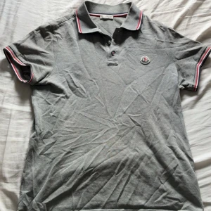 Moncler piké - Snygg grå pikétröja från Moncler med klassisk krage och knäppning framtill. Röd och vit rand på krage och ärmslut ger en sportig vibe. Broderad Moncler-logga på bröstet. Perfekt till jeans eller shorts.