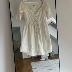 Vacker sommarklänning  - Supersöt vit kortklänning från Spira med broderade spetsdetaljer, knappar framtill och knyt i midjan. Klänningen har v-ringning och halvlånga ärmar. Storlek S.