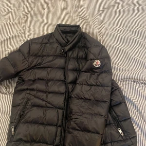 Svart Moncler dunjacka herr - Säljer en svart dunjacka från Moncler med klassisk logga på ärmen. Jackan har hög krage, dragkedja framtill och två fickor med dragkedja. Perfekt för kalla dagar och riktigt snygg till streetwear. Fylld med dun och har en clean, quiltad look. Storlek är S eller M