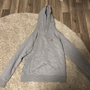 Grå hoodie från GANT med logga - Grå hoodie från GANT med broderad logga och sköld på bröstet. Klassisk känguruficka framtill och stor huva. Mjuk och skön i bomullsmix, perfekt för chill dagar. Ribbade muddar vid ärmslut och nederkant.