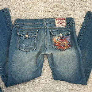 True Religion jeans med drakbrodyr - Blå jeans från True Religion med snyggt sliten tvätt och broderad drake i färg på bakfickan. Klassiska kontrastsömmar, utsvängda ben och ikoniska fickdetaljer. Jeansen är lågmidjade och bootcut i storlek 30, s/m