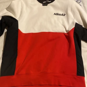 Nike Air röd och vit sweatshirt - Nike Air sweatshirt i färgblockad design med rött, vitt och svarta detaljer. Tröjan har lång ärm, rund hals och tryckt logga på bröstet. Perfekt för en sportig och avslappnad stil.