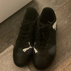 Svarta Puma fotbollsskor med vit logga - Snygga svarta fotbollsskor från Puma med vit logga på sidan och ovansidan. Skorna har snörning och en låg profil, perfekt för snabba rörelser på planen. Tillverkade i syntetmaterial med mönstrad yta för extra grepp och stil.