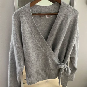 Grå omlottkofta från H&M - Mysig grå kofta från H&M i omlottmodell med knyt i sidan. Koftan har lång ärm och bred ribbad kant nedtill. Perfekt för lager-på-lager och chill dagar. Superenkel att matcha med jeans eller kjol.