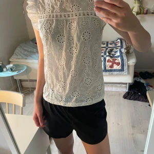 Vit blus med broderade blommor - Supersöt vit blus i bomull med broderade blommor och hålmönster. Blusen har volangärmar, rund halsringning och knapp i nacken. Perfekt för en fräsch och somrig look med romantiska detaljer.