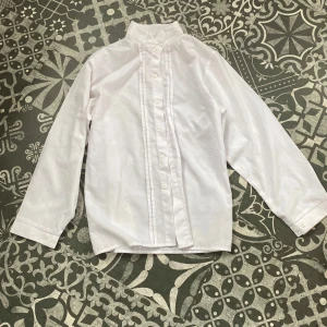 Vit henley-skjorta med ståkrage - Klassisk vit henley-skjorta med ståkrage och knappar framtill. Skjortan har lång ärm och diskreta veck längs knappslån för en stilren look. Aldrig använd ☀️