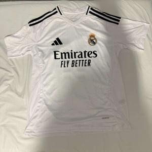 Real Madrid vit matchtröja Adidas - Säljer en vit Real Madrid fotbollströja från Adidas med svarta detaljer och klubbmärke på bröstet. Tröjan har korta ärmar, svarta ränder på axlarna och tryck med 'Emirates Fly Better' samt namn och nummer 11 på ryggen. Tillverkad i lätt och ventilerande material.