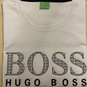 Vit t-shirt från Hugo Boss med logga - Helt ny och oanvänd vit t-shirt från Hugo Boss med stor BOSS-logga framtill och tryckt mönster i bokstäverna. Klassisk rund halsringning och korta ärmar. Skön och stilren modell i mjuk bomull, perfekt för dig som gillar snygga loggor. Står XL men är liten i storlek och passar M/L