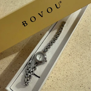Silverfärgad klocka från Bovou - Stilren och nätt klocka från Bovou med rund urtavla och länkarmband i silverfärgad metall. Urtavlan är minimalistisk med diskreta visare och Bovou-logga. Perfekt accessoar för dig som gillar en clean look och snygga detaljer. I stort sett så ser den ny ut, har ej några defekter och funkar bra. Det tillkommer även kvitto och tre extra länkar!