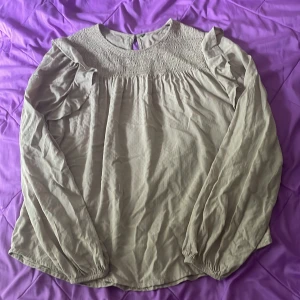 Beige blus med smock och volang - Supersöt beige blus med smock upptill och volangdetaljer på axlarna. Långärmad med resår vid ärmslut och diskret strukturmönster i tyget. Rund halsringning och knapp i nacken. Perfekt för dig som gillar en romantisk och chill stil.