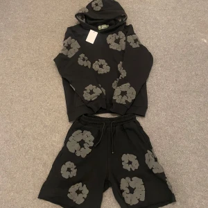 Denim tears Set - Svarta shorts och hoodie med unikt grått blommönster och avslappnad passform. Resår i midjan med snörning för extra komfort. Perfekta för en chill och trendig stil under varmare dagar.