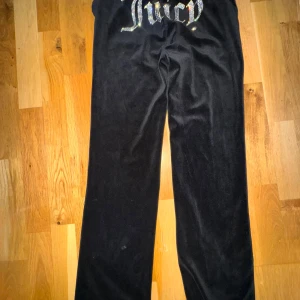 Svarta velourbyxor Juicy Couture XS - Svarta mjukisbyxor från Juicy Couture i velour med glittrande strasstenar som bildar texten 'Juicy' på baksidan. Byxorna har normal passform, snörning i midjan och en liten strasstenad 'J' framtill. Perfekta för en chill och trendig look.
