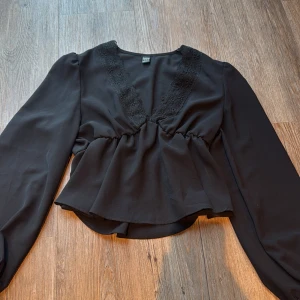 Svart blus med spets från Droimp - Svart blus från Droimp med lång ärm och v-ringad hals som är dekorerad med snygg spets. Modellen har en lätt peplum-effekt med rynk vid midjan och ett flowigt fall. Perfekt för dig som gillar stilrena detaljer och vill ha en feminin touch.