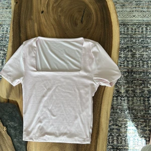 Ljusrosa topp med fyrkantig hals - Snygg ljusrosa topp med korta ärmar och en modern fyrkantig halsringning. Toppens stilrena design gör den enkel att matcha med olika outfits. Materialet känns mjukt och stretchigt, perfekt för dig som gillar bekväma och trendiga plagg.
