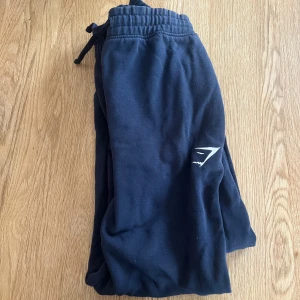 Mörkblå mjukisbyxor från Gymshark - Snygga mörkblå mjukisbyxor från Gymshark med logga på benet. Byxorna har snörning i midjan och är gjorda i mjukt bomullsmaterial. Passformen är mindre än vanligt och de är perfekta för chill eller träning. Enkla att matcha med hoodies eller t-shirts.