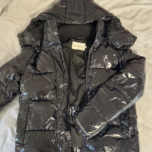 Svart pufferjacka från Calvin Klein Jeans - Snygg svart pufferjacka från Calvin Klein Jeans med glansig finish och logga på bröstet. Jackan har avtagbar huva, dragkedja och stora fickor framtill. Väldigt varm så perfekt inför hösten och vintern💕Använd sparsamt en vinter och ser ut som ny! köpt för ungefär 2200kr.