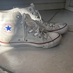 Converse Chuck Taylor vita high tops - Säljer ett par klassiska Converse Chuck Taylor All Star high tops i vit canvas med röd och blå rand på sulan. Skorna har snörning, rund tå och platt sula. Ikonisk logga på sidan och metallöglor för snören. Perfekta för dig som gillar retrostil och streetwear.