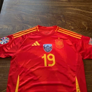 Spaniens landslagströja Lamine Yamal - Säljer en röd Spanien fotbollströja med gula detaljer från Adidas, nummer 19 och Lamine Yamal på ryggen. Tröjan har korta ärmar, rund hals och officiella landslagsmärken samt turneringspatch på ärmen. Aldrig använd men utan prislappen! 