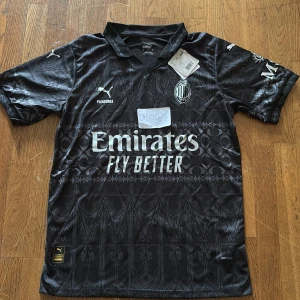 Svart AC Milan x Puma fotbollströja - AC Milan x Puma fotbollströja i svart med detaljerat grafiskt mönster och vita tryck. Kortärmad med krage, tillverkad i lätt DryCell-material. Loggor från Puma, Pleasures och AC Milan samt Emirates Fly Better på bröstet. Perfekt för dig som vill sticka ut på planen.