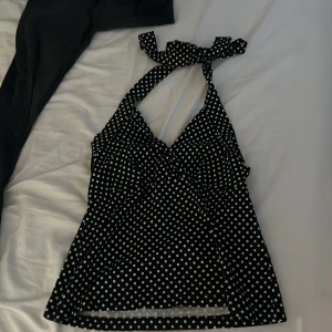 Svart prickig halterneck topp Bodyflirt - Svart halterneck topp från Bodyflirt med vita prickar och snygg knytning i nacken. Toppen har en feminin v-ringad form och är lätt utsvängd nedtill. Perfekt för dig som gillar retrovibbar och vill sticka ut med ett coolt mönster.