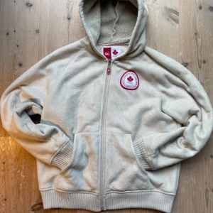 Beige fleece jacka från HBC Canada - Säljer en beige jacka i mjuk fleece från HBC med Canadian Olympic Team patch på bröstet och stor röd CANADA-text på ryggen. Tröjan har huva, dragkedja och två fickor fram. Riktigt snygg! Köpt på betong retro. Har ett litet märke (se bild). Står storlek L men sitter som en M. Skriv vid intresse!