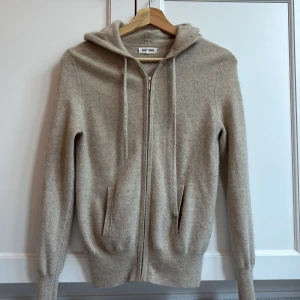 Beige hoodie i kashmir från Soft Goat - Mjuk och lyxig beige hoodie från Soft Goat i kashmir. Tröjan har dragkedja framtill, snörning vid huvan och två fickor. Långärmad med ribbade muddar och en clean, oversized look. Perfekt för dig som gillar stilren och comfy vibe.