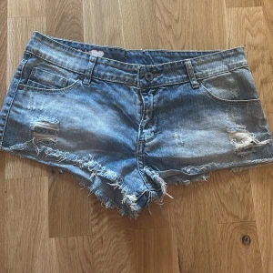 Lowaist Blå jeansshorts med slitningar - Snygga blå jeansshorts med fransiga kanter och slitna detaljer framtill. Klassisk femficksmodell med knapp och dragkedja. Broderad rosa hjärta och logga baktill ger en extra touch. Perfekta för varma dagar och en avslappnad stil.
