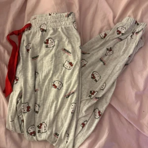 Hello Kitty julmönstrade mjukisbyxor - Supermysiga ljusgrå mjukisbyxor med Hello Kitty-motiv och juliga detaljer som snöflingor och texten 'Merry Christmas'. Byxorna har röd snörning i midjan och elastisk linning för extra komfort. Perfekta för chill dagar hemma eller julmys! Finns i storlek M och S