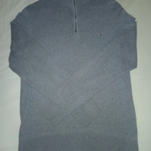 Grå half zip tröja Tommy Hilfiger - Säljer en stilren grå långärmad tröja från Tommy Hilfiger med half zip och liten logga på bröstet. Tröjan är gjord i mjuk bomull och kashmirblandning, har ribbade muddar och klassisk krage. Perfekt för dig som gillar enkel och snygg design.