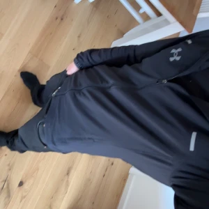 Svart vindjacka från Under Armour - Snygg svart vindjacka från Under Armour med huva och hel dragkedja framtill. Jackan har vita logodetaljer på bröstet och reflekterande inslag vid axlarna. Långärmad modell med ribbade muddar där Under Armour är broderat. Perfekt för aktiva dagar.