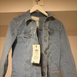 Ljusblå jeansjacka från ONLY XS - Snygg ljusblå jeansjacka från ONLY i klassisk modell med raka linjer och silverfärgade knappar framtill. Jackan har två bröstfickor med knapp och lång ärm. Perfekt att slänga över en fin topp undertill! Den är helt ny med prislapp kvar!