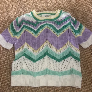Pastellfärgad stickad tröja - Säljer en kortärmad stickad tröja från byclara med zigzag-mönster i pastellfärger som lila, mintgrön, vit och lite gult. Tröjan har rund hals och är luftig med hålstickat material, perfekt för vår och sommar. Superfin att styla med jeans eller kjol!