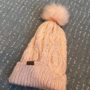 Stickad rosa mössa med pälsboll från H&M - Säljer en supermysig stickad mössa i ljusrosa från H&M, med en fluffig pälsboll på toppen. Mössan har grovstickat flätmönster och uppvikt kant med en liten etikett där det står 'Adventures are forever'. Perfekt för kalla dagar och riktigt snygg till vinterlooken.